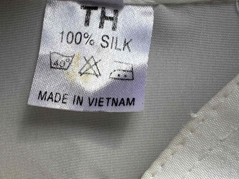 Op de afbeelding: Een wit stoffen label met de tekst "TH", "100% SILK" en "MADE IN VIETNAM" in het zwart. Het label bevat ook wassymbolen. Het label is op een lichtgrijze stof genaaid.