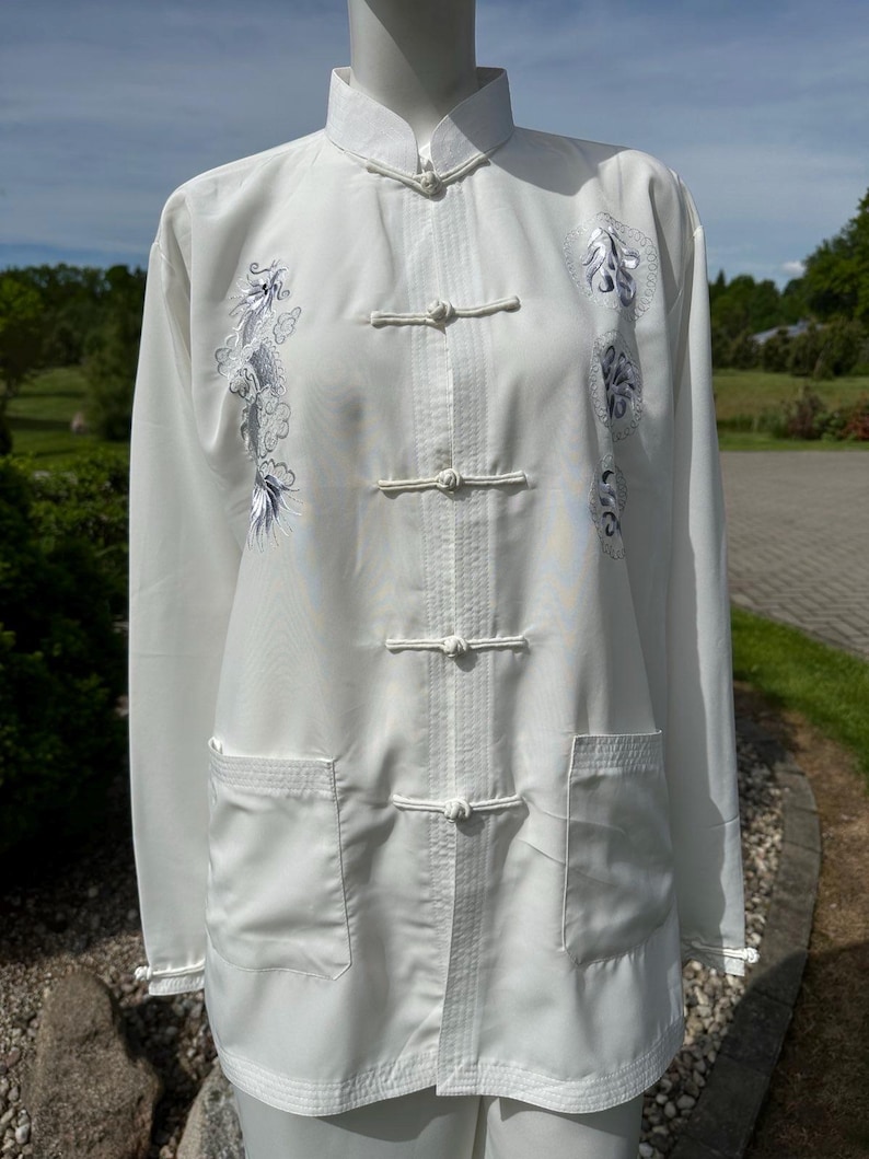Op de afbeelding: Witte vechtsportuniform met een Chinese kraag en kikker sluitingen. Het uniform heeft geborduurde draken- en bloemmotieven in blauw- en grijstinten. Twee voorzakken zijn zichtbaar.
