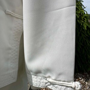 Op de afbeelding: Close-up van een wit kledingstuk, mogelijk een jas of top, met een gladde, matte textuur. De mouw heeft een decoratieve manchet met een geknoopte sluiting. De stiksels zijn zichtbaar en het algehele ontwerp is eenvoudig en elegant.