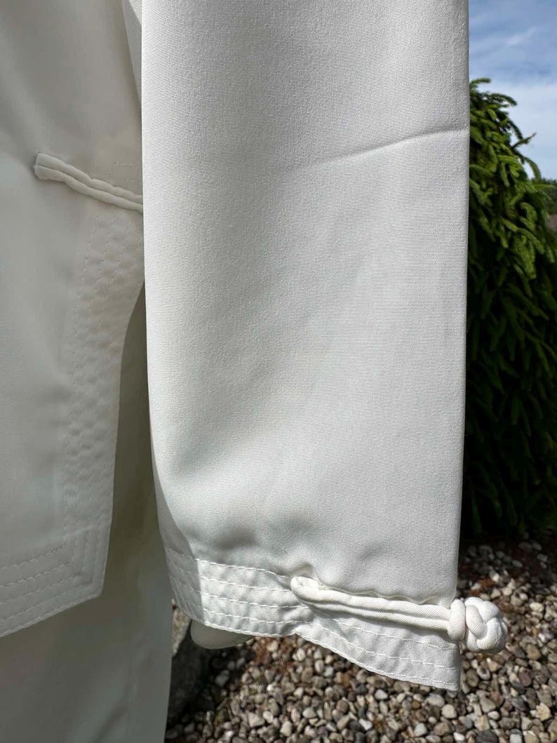 Op de afbeelding: Close-up van een wit kledingstuk, mogelijk een jas of top, met een gladde, matte textuur. De mouw heeft een decoratieve manchet met een geknoopte sluiting. De stiksels zijn zichtbaar en het algehele ontwerp is eenvoudig en elegant.