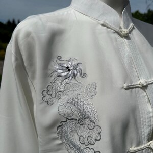 Op de afbeelding: Witte vechtsportuniform met een mandarijnkraag en kikkerknopen. Het uniform heeft een geborduurd zilveren drakenontwerp op de voorkant. De top met lange mouwen is gemaakt van een gladde, mogelijk zijdeachtige stof.