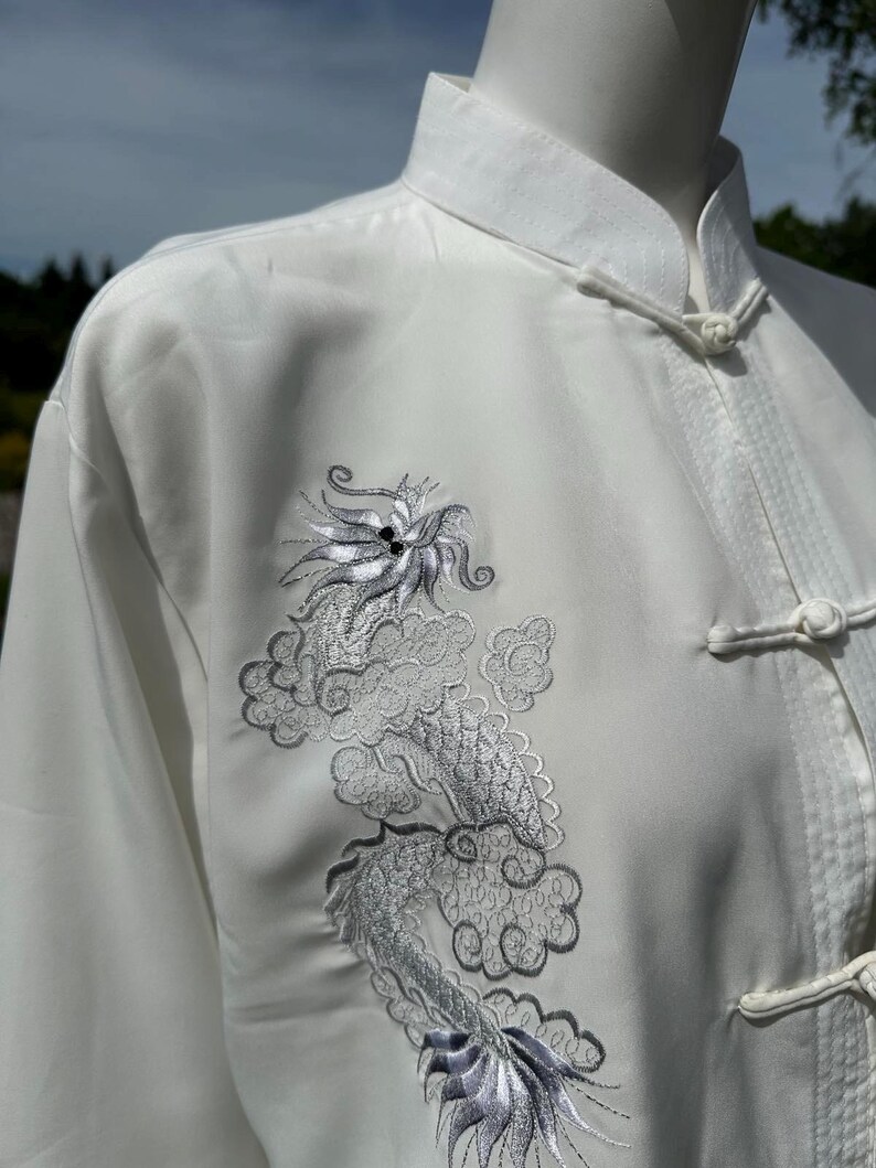 Op de afbeelding: Witte vechtsportuniform met een mandarijnkraag en kikkerknopen. Het uniform heeft een geborduurd zilveren drakenontwerp op de voorkant. De top met lange mouwen is gemaakt van een gladde, mogelijk zijdeachtige stof.