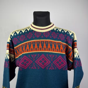 May include: A teal sweater with a cream-colored collar and cuffs. The sweater features a geometric pattern in orange, pink, and cream. The sleeves have a similar design. The sweater is displayed on a black mannequin.