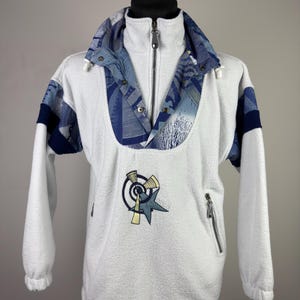 May include: White fleece pullover with a blue and white patterned collar and shoulder accents. Features a full zipper, a small embroidered graphic, and a zippered pocket. The sleeves are long and the cuffs are elastic.