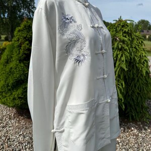 Op de afbeelding: Wit shirt met lange mouwen, een Chinese kraag en kikkerknopen. Het shirt heeft een gedetailleerd geborduurd drakendesign in grijstinten en paars op de voorkant. Het shirt heeft twee voorzakken en is gemaakt van een soepele, vloeiende stof.