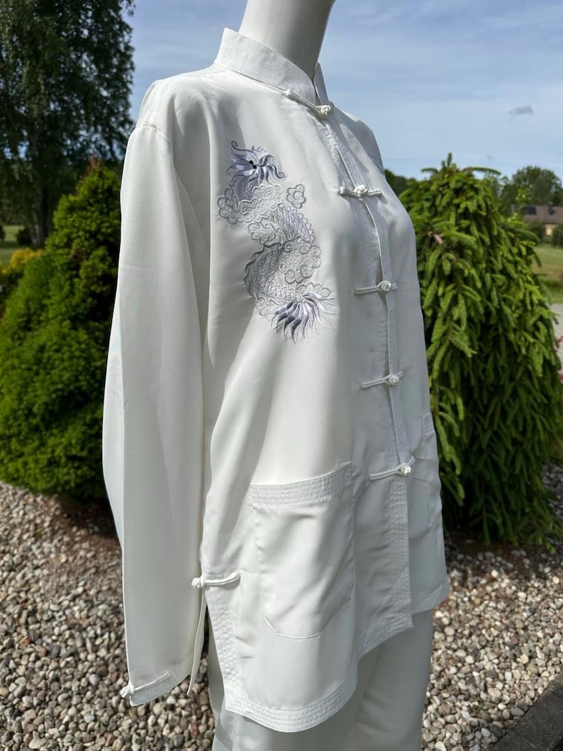 Op de afbeelding: Wit shirt met lange mouwen, een Chinese kraag en kikkerknopen. Het shirt heeft een gedetailleerd geborduurd drakendesign in grijstinten en paars op de voorkant. Het shirt heeft twee voorzakken en is gemaakt van een soepele, vloeiende stof.