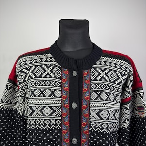 May include: A black, white, and red patterned cardigan sweater with a button-up front. The sweater features a geometric design with floral accents and a round neckline. The sleeves and shoulders have red accents. The sweater is displayed on a black mannequin.