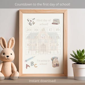 Könnte beinhalten: Ein gerahmter Druck mit einem Countdown-Kalender für den ersten Schultag. Der Druck enthält Illustrationen von Schulmaterialien und den Text "Countdown to the first day of school!" und "I'm going to school!". Ein Plüschhase und eine Topf-Sukkulente befinden sich neben dem Rahmen.