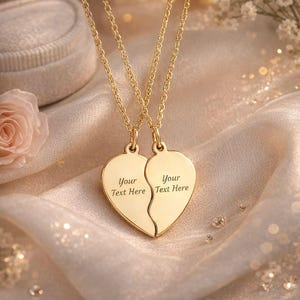 Peut inclure: Deux colliers pendentifs en forme de cœur dorés, chacun gravé de "Your Text Here", reliés à de fines chaînes. Les pendentifs sont fendus pour former un cœur complet. Le fond présente un tissu satiné et une rose.