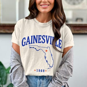 Puede incluir: Camiseta color crema con la palabra "GAINESVILLE" en texto azul arqueado. Debajo, un contorno azul de Florida con una estrella roja. El año "1869" está impreso debajo del mapa en azul.