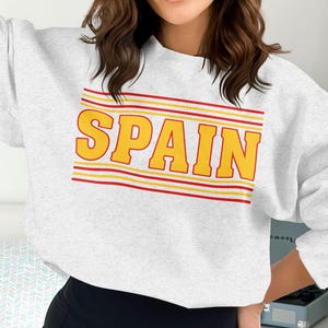 May include: A light gray sweatshirt with the word "SPAIN" in yellow block letters. The word is outlined with red and yellow stripes. The sweatshirt has long sleeves and a crew neck.