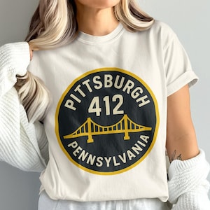 Peut inclure: T-shirt crème avec un motif circulaire. Le motif comprend le texte "PITTSBURGH 412 PENNSYLVANIA" autour d'un graphique de pont jaune. Le t-shirt a des manches courtes et une coupe décontractée.