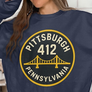 May include: Navy blue sweatshirt featuring a circular graphic with the text "PITTSBURGH 412 PENNSYLVANIA". The graphic includes a stylized bridge illustration in yellow, set against a black background with a yellow border. A casual top.