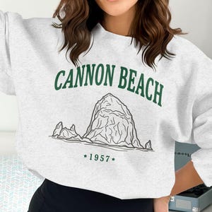 Peut inclure: Un sweat-shirt gris clair avec l'inscription "CANNON BEACH" en vert au-dessus d'une illustration en noir et blanc d'une formation rocheuse. Le texte "*1957*" est en dessous.