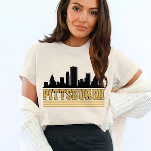 May include: A cream-coloured short-sleeve t-shirt featuring a black silhouette of the Pittsburgh skyline. Below the skyline, the word PITTSBURGH is printed in a bold, gold-coloured font, underlined by three horizontal gold-coloured lines.