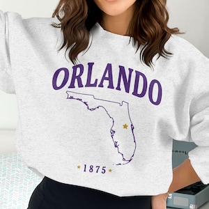 May include: A light grey sweatshirt featuring the word "ORLANDO" in purple lettering. Below is a purple outline of Florida with a star. The year "1875" is below the map in purple with stars.