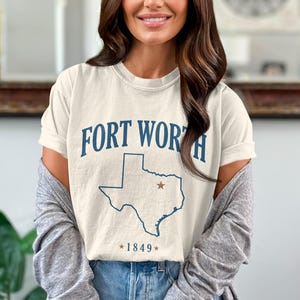 May include: Cream-colored t-shirt featuring the text "FORT WORTH" in navy blue, along with a navy blue outline of the state of Texas and a star. The year "1849" is printed below the Texas outline. The shirt is worn by a person.