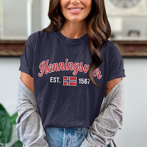 May include: A dark navy blue short-sleeved t-shirt featuring the white text Henningsvær in a cursive font above the text EST. 1567 with a small rectangular Norwegian flag graphic centered between the numbers on the front of the garment.