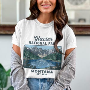 May include: White t-shirt featuring a scenic illustration of Glacier National Park in Montana. The design includes a lake, mountains, and trees, with the text "Glacier National Park Montana EST. 1910". The shirt is a classic crew neck.