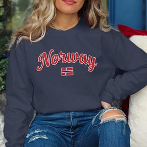 May include: A dark navy blue crewneck sweatshirt featuring the word Norway written in a red cursive font with a white outline. Below the text is a small rectangular flag of Norway with a red background, a white cross, and a blue cross.