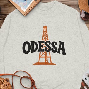 Może przedstawiać: Jasnoszara bluza z napisem "ODESSA" w kolorze czarnym i pomarańczową grafiką wieży wiertniczej. Bluza ma okrągły dekolt. Zdjęcie znajduje się na drewnianej powierzchni z sandałami, kapeluszem i aparatem.