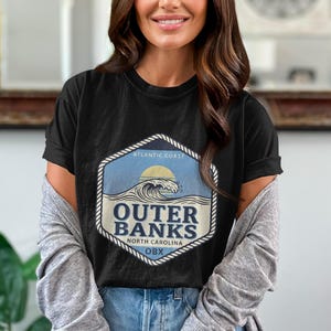 May include: A black short-sleeve t-shirt featuring a hexagonal graphic with a rope border. The design includes a blue ocean wave, a yellow sun, and the text "ATLANTIC COAST," "OUTER BANKS," "NORTH CAROLINA," and "OBX" in dark blue lettering.