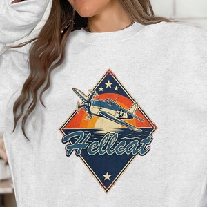 May include: A light gray sweatshirt featuring a vintage-style graphic of a blue and white airplane with the word "Hellcat" in a diamond-shaped design. The graphic includes a sunset and stars.