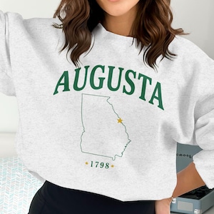 Può includere: Felpa grigio chiaro con la scritta "AUGUSTA" in lettere verdi. Sotto, un contorno verde dello stato della Georgia con una stella dorata. Il numero "1798" è sotto il contorno dello stato.