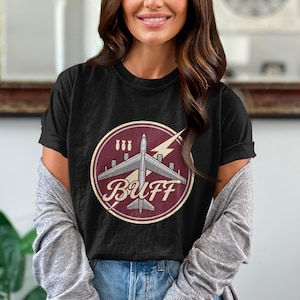 May include: A black short-sleeve t-shirt featuring a circular graphic on the chest. The design displays a gray military bomber aircraft, three small bomb icons, a yellow lightning bolt, and the text BUFF in a stylized cream-colored font.