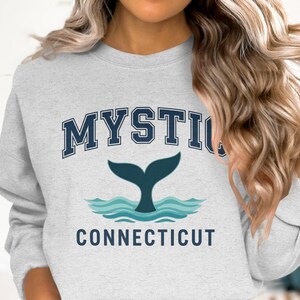 Può includere: Una felpa grigio chiaro con la scritta "MYSTIC" in lettere maiuscole blu navy sopra un disegno di coda di balena e la scritta "CONNECTICUT" sotto. La coda di balena è blu navy e le onde sono azzurre.