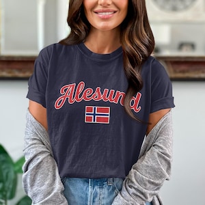 May include: A navy blue short-sleeve t-shirt featuring the word Alesund in a red cursive font with a white outline. Below the text is a rectangular graphic of the Norwegian flag, which consists of a red background with a blue cross outlined in white.