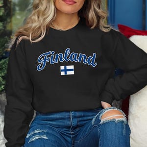 May include: A black long-sleeve crewneck sweatshirt featuring the word Finland written in a blue script font with a white outline, positioned above a small graphic of the national flag of Finland, which consists of a blue cross on a white field.