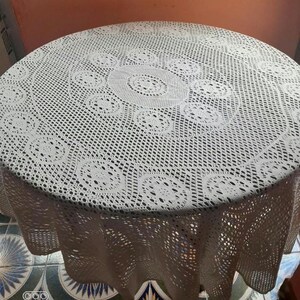 May include: A cream-colored, round crocheted tablecloth. The intricate lace design features a repeating pattern of circular motifs. The tablecloth is draped over a round table, showcasing the detailed craftsmanship.