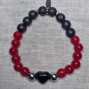 May include: A bracelet with red and black beads, a black heart-shaped bead, and silver-coloured beads. The beads are strung on an elastic cord. The bracelet has a small metal tag. The beads are arranged in an alternating pattern.