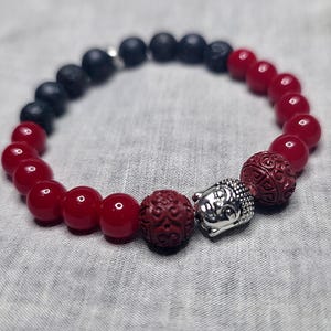 May include: A bracelet with red and black beads. The bracelet has two carved red beads and a silver-coloured Buddha head charm. The beads are strung on an elastic cord. The beads are measured in centimetres.