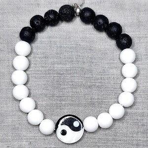 May include: A bracelet with black and white beads, featuring a yin-yang symbol. The bracelet alternates between black lava rock beads and white round beads. A small, clear quartz charm hangs from the clasp. The bracelet is designed for everyday wear.