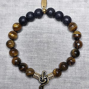 May include: A beaded bracelet with alternating black and brown tiger eye beads. The bracelet features a gold-coloured treble clef charm and a small gold-coloured tag. The beads are approximately 0.8 cm in diameter.