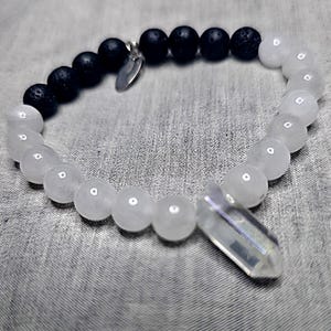 May include: A bracelet with black lava stone beads and white moonstone beads. A clear quartz crystal pendant is attached. A small silver charm is also present.