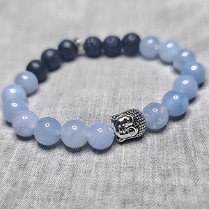 Peut inclure: Bracelet de perles bleues claires et noires, orné d'un charm en forme de tête de Bouddha argentée. Les perles sont enfilées sur un élastique. Le bracelet est présenté sur une surface grise. Les perles mesurent environ 0,8 cm de diamètre.