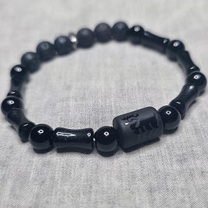 May include: Black beaded bracelet with a combination of round and cylindrical beads. The bracelet has a central rectangular bead with an etched design. The beads are strung on an elastic band, creating a flexible and stylish accessory.