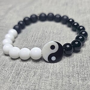 May include: A bracelet with a yin-yang symbol at its centre. The bracelet is made of white and black beads, with a larger yin-yang bead. The beads are strung together on an elastic band. The bracelet is displayed on a grey surface.