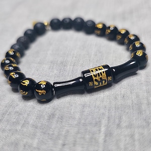 Op de afbeelding: Zwarte kralenarmband met goudkleurige accenten. De armband heeft ronde kralen met gouden symbolen en een centrale zwarte cilindrische kraal met gouden details. De armband ligt op een grijze ondergrond.