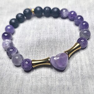May include: A bracelet with a combination of dark black and light purple beads. The bracelet features a heart-shaped amethyst stone in the centre, connected by gold-coloured metal tubes. The beads are round and smooth, creating a stylish accessory.