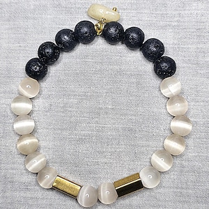 May include: A bracelet with black lava rock beads and white cat's eye beads. It features gold-coloured metal bars and a small, off-white charm. The bracelet is displayed on a grey fabric surface.