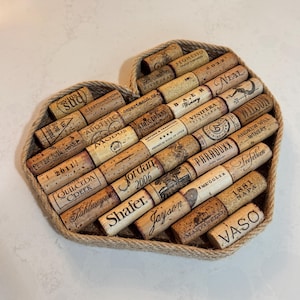May include: A heart-shaped tray filled with assorted wine corks. The corks display various winery names and dates, arranged within a woven rope border. The tray sits on a white surface.