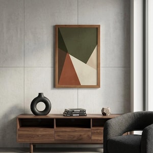 May include: A framed abstract art print featuring geometric shapes in shades of green, brown, and beige. The artwork is displayed in a wooden frame. A black vase, books, and a decorative object sit on a wooden cabinet below.