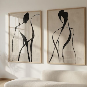 Op de afbeelding: Twee ingelijste wandkunstwerken met abstracte zwarte inkttekeningen van menselijke figuren op een neutrale beige achtergrond. Elk rechthoekig stuk is gevat in een lichtgekleurde houten lijst en toont vloeiende, minimalistische lijnen.