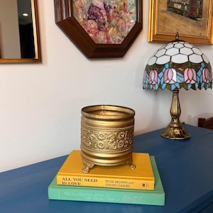 May include: A gold-colored, ornate candle holder with floral detailing sits atop two books. The books are titled "ALL YOU NEED IS LOVE" and "THE ILLUSTRATED GUIDE TO THE COLLECTIBLES OF Coca-Cola." A stained glass lamp and framed art pieces are in the background.