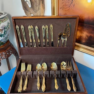 Puede incluir: Un juego de cubiertos dorado vintage exhibido en un estuche de madera con forro de terciopelo marrón. El juego incluye tenedores, cucharas, cuchillos y utensilios para servir. Los mangos presentan un diseño floral ornamentado.
