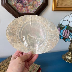 Puede incluir: Plato ovalado de nácar iridiscente con diseños florales y de volutas grabados. El plato tiene un área central lisa y un borde decorativo. El plato se sostiene frente a una superficie azul, con libros y obras de arte en el fondo.